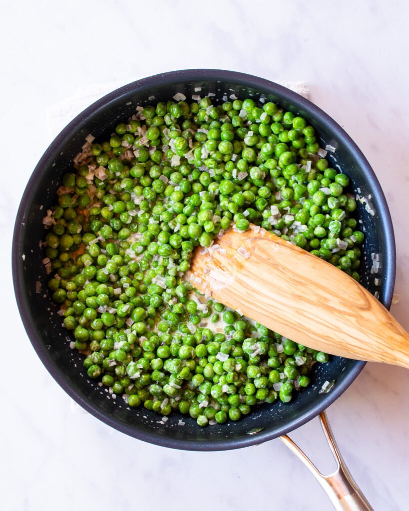A wooden spoon in a skillet with green peas.