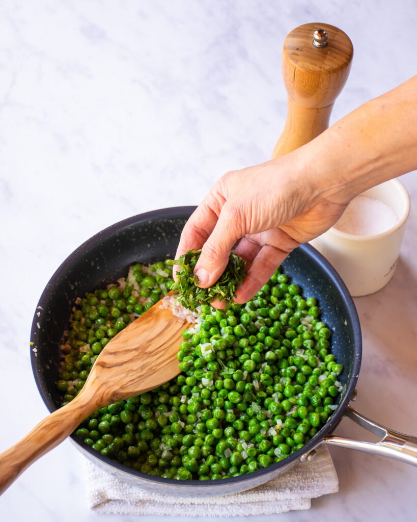 A hand adding mint to a skillet of peas.