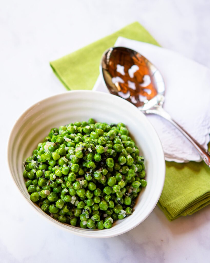 Minted peas in a ribbed white bowl with a serving spoon and green and white napkins.