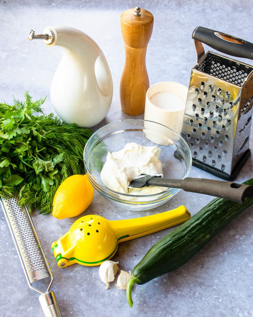 Ingredients on a countertop - lemon and lemon squeezer, cucumber, fresh dill and parsley, salt, pepper, olive oil, yogurt and a box grater.