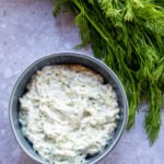A blue bowl of Tzatziki sauce with fresh dill next to it.