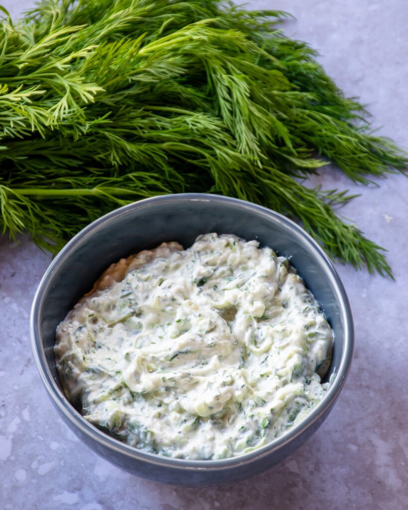 A blue bowl of Tzatziki sauce with fresh dill next to it.
