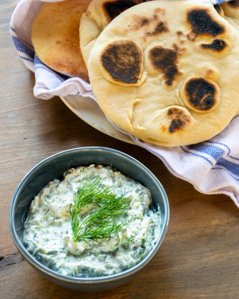 Tzatziki sauce in a blue bowl with a plate of homemade pita bread.