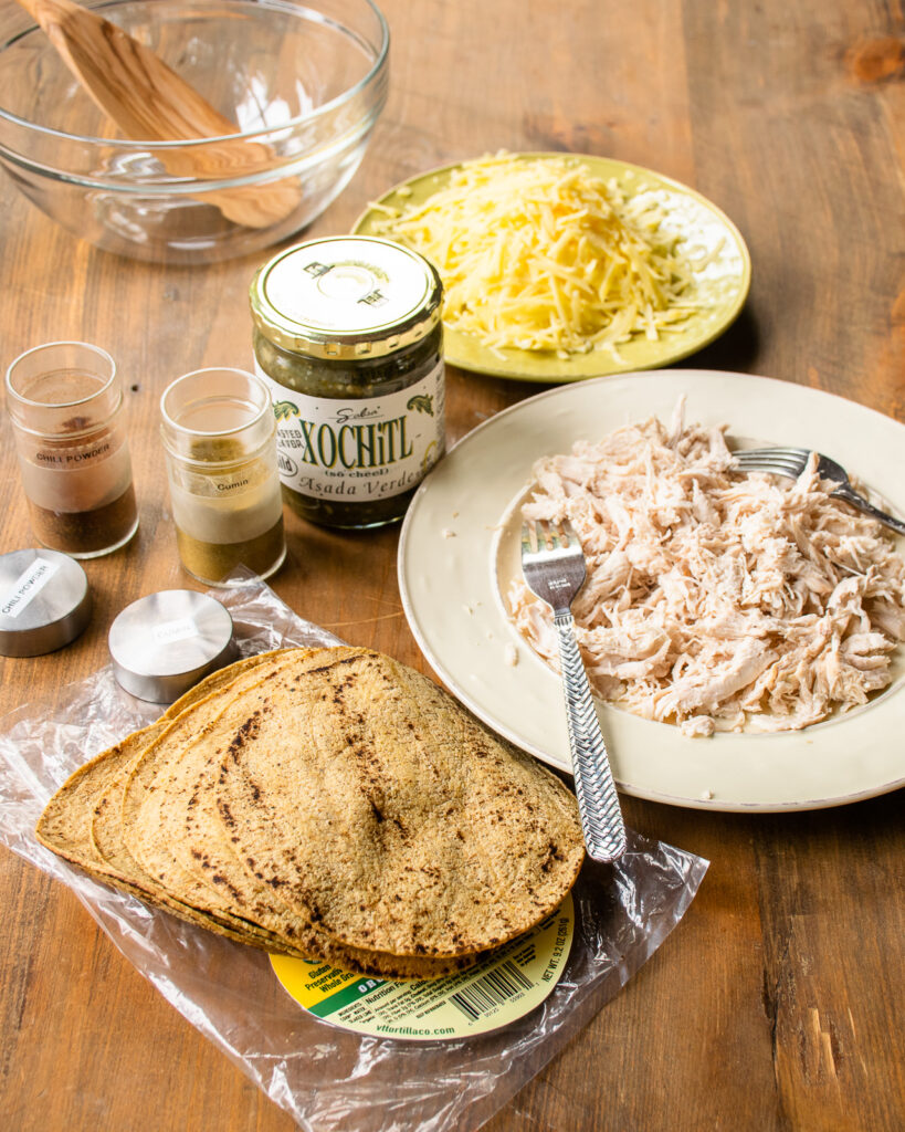 Ingredients on a wooden table - shredded chicken, corn tortillas, green salsa, spices and cheese.