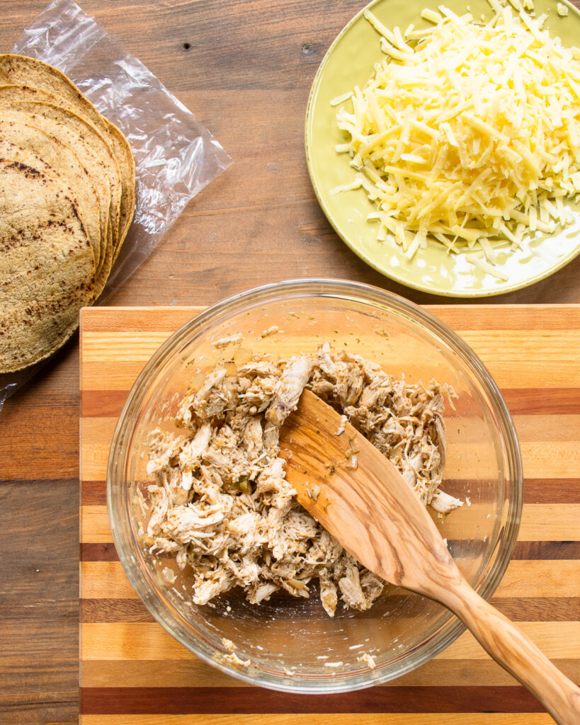 Chicken taquitos filling mixed in a bowl with tortillas and cheese near by.