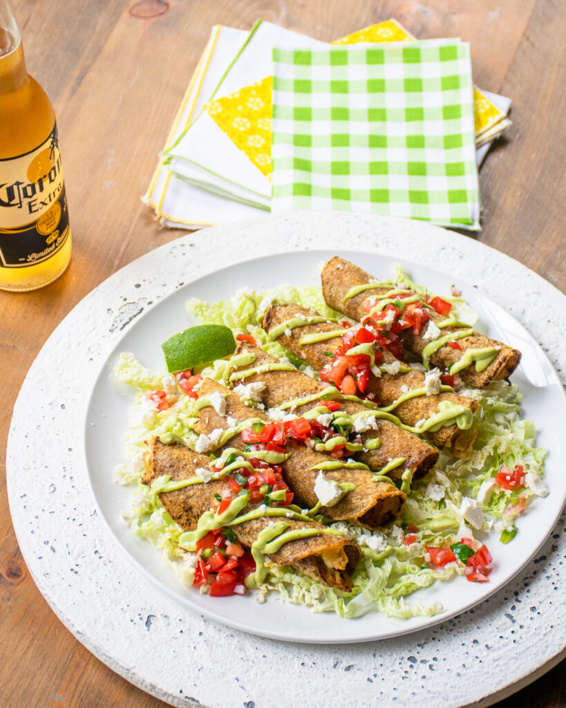 Chicken and green chili taquitos on a white plate with a green checkered napkin and a bottle of beer.