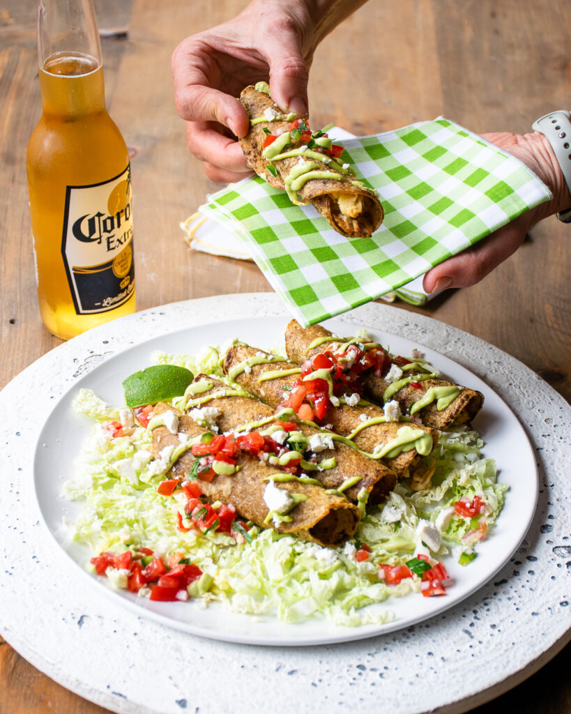 A hand picking up a taquito from a white plate with a green checkered napkin and a bottle of beer.