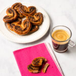 Chocolate Palmiers on a white plate with one palmier on a pink beverage napkin with an espresso.