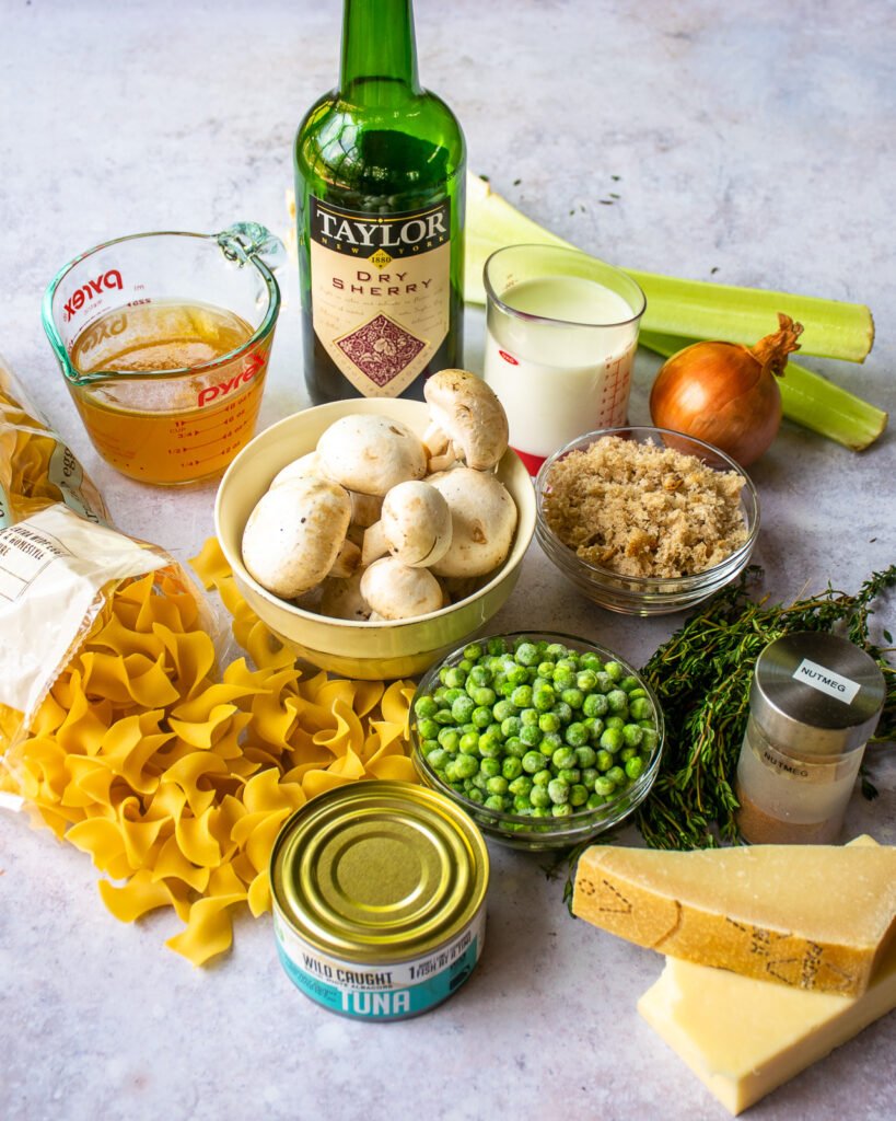 Ingredients on a countertop - a can of tuna, noodles, peas, mushrooms, cheeses, breadcrumbs, vegetables, milk, stock and sherry.