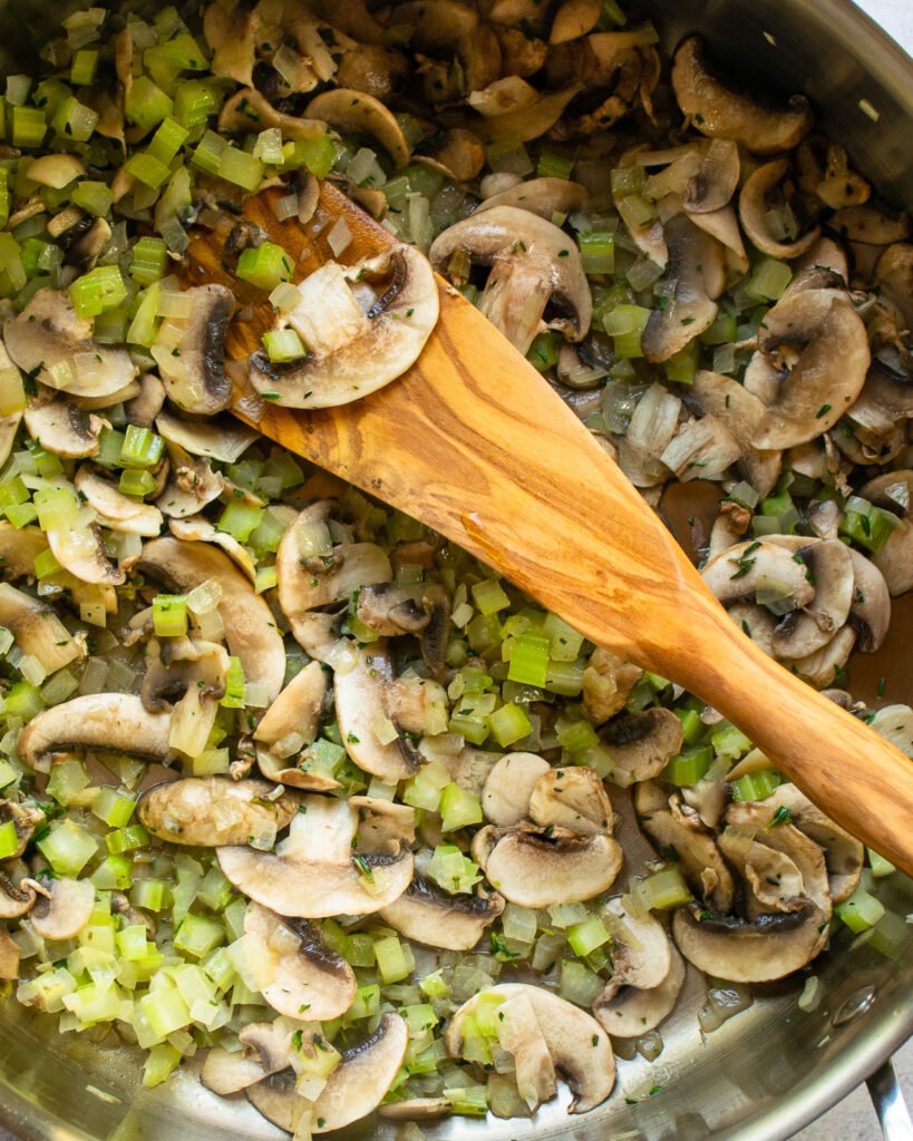 Onion, celery and mushrooms cooking in a pan with a wooden spoon.