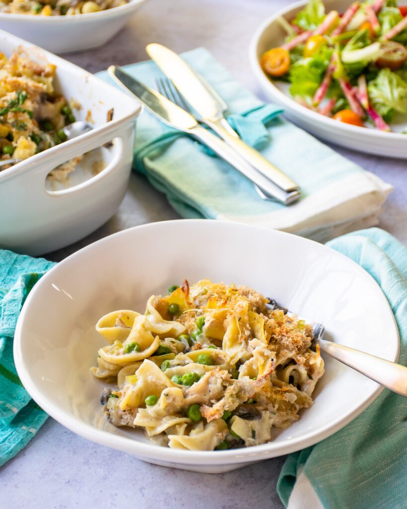 A serving of tuna noodle casserole with salad, a casserole dish and turquoise napkins in the background.