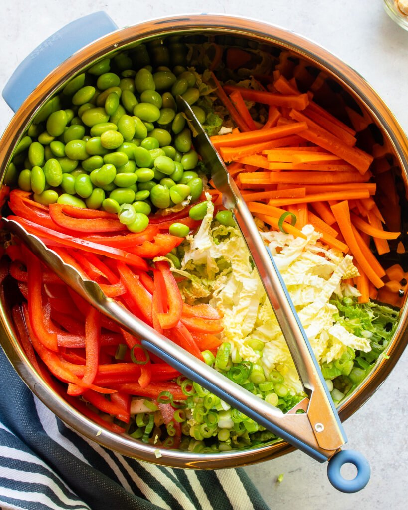Ingredients for Asian slaw in a large bowl with a pair of tongs.