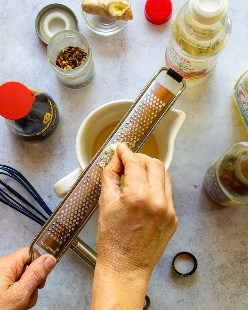 Garlic being grated into a ceramic measuring glass with ingredients around.