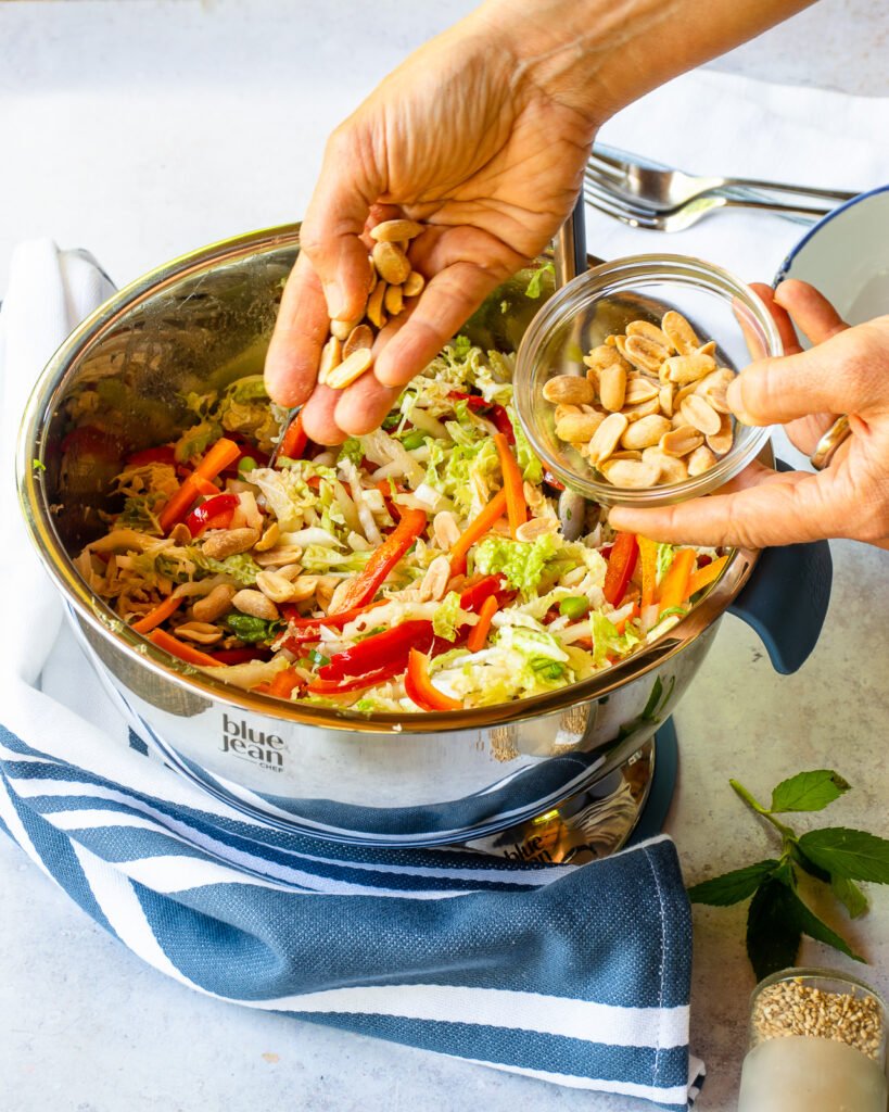 Hands scattering peanuts into a bowl of Asian slaw with a blue and white striped towel around the bowl.