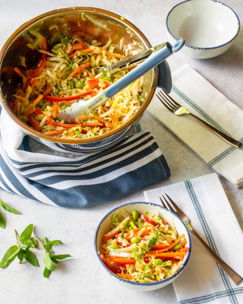 A big bowl of Asian slaw with tongs coming out of the bowl, next to a smaller white bowl of slaw and a white and blue striped napkin.