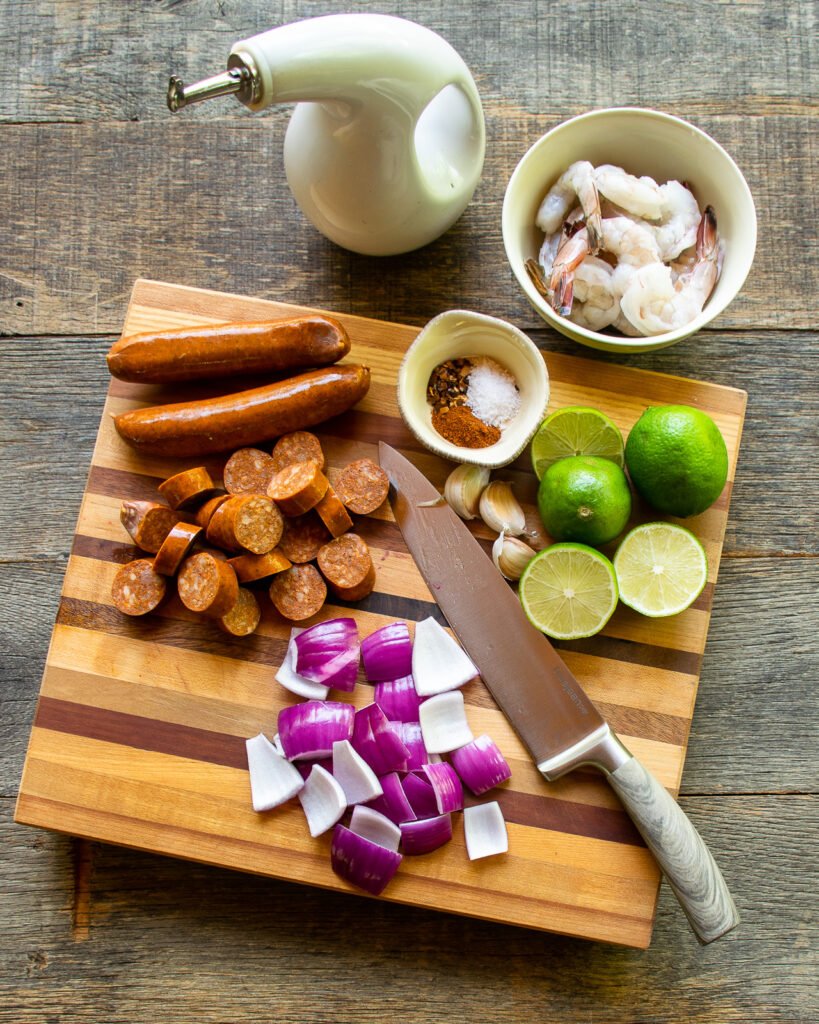 Ingredients on a wooden cutting board - red onion, sliced chorizo, limes, spices, a bowl of shrimp and a bottle of olive oil.