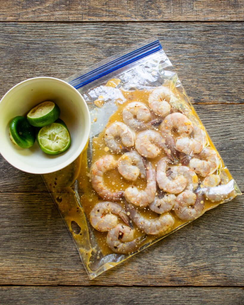 Shrimp marinating in a plastic bag with a small bowl of lime rinds next to it.