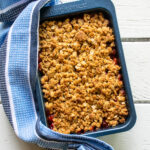 Strawberry Rhubarb crisp in a blue baking dish with a blue striped towel on a white wooden table.