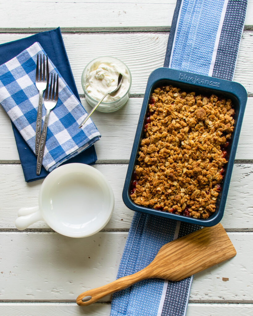 Strawberry Rhubarb Crisp in a blue pan on a blue striped towel with white bowls and blue checkered napkins all on a white wooden table.