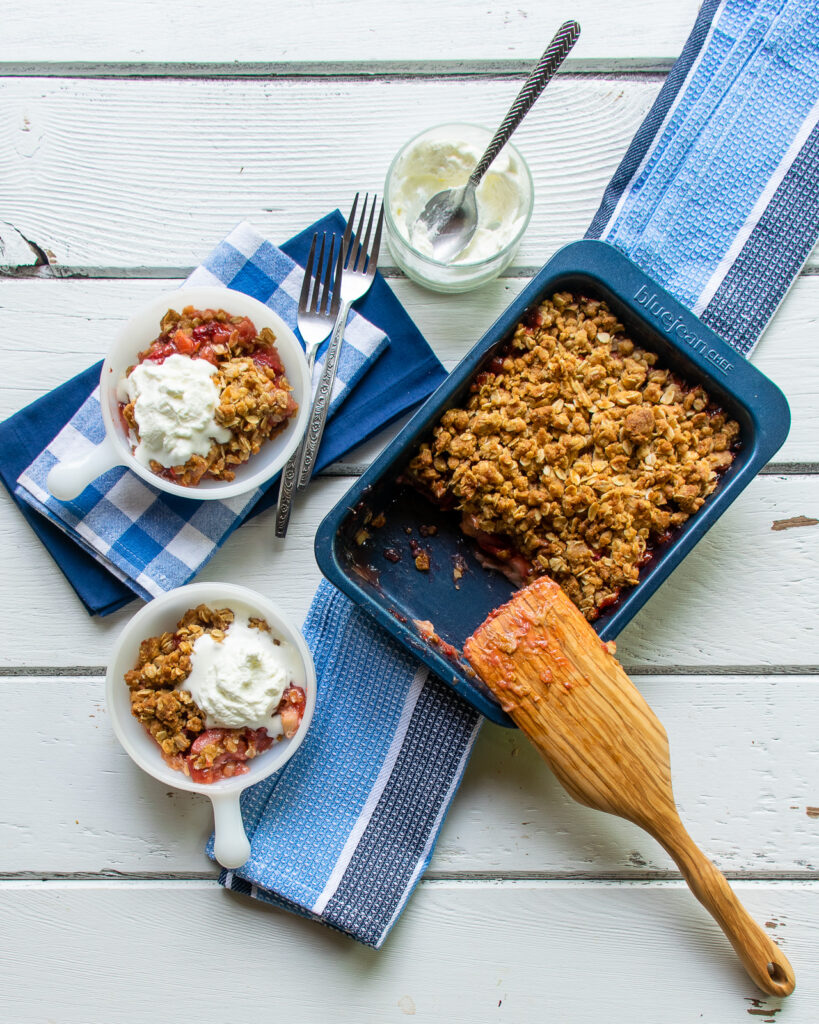Strawberry Rhubarb Crisp in a blue pan on a blue striped towel with white bowls and blue checkered napkins all on a white wooden table.