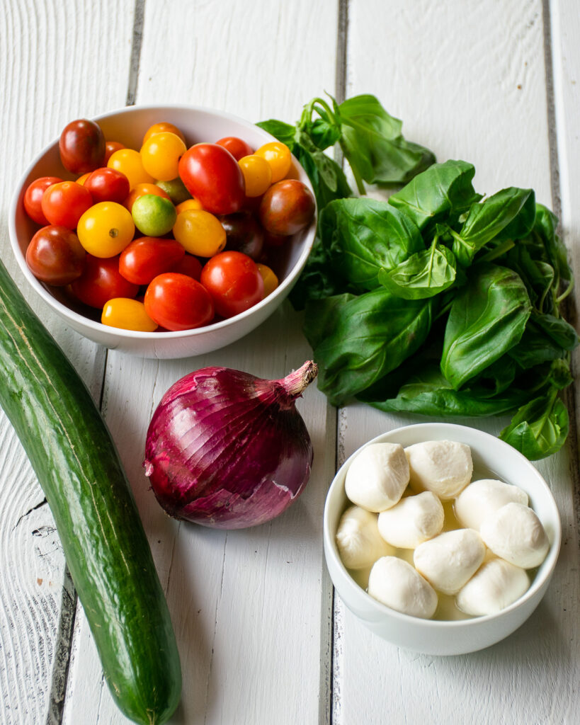 A bowl of colored tomatoes with a cucumber, red onion, basil and fresh mozzarella balls on a white wooden table.