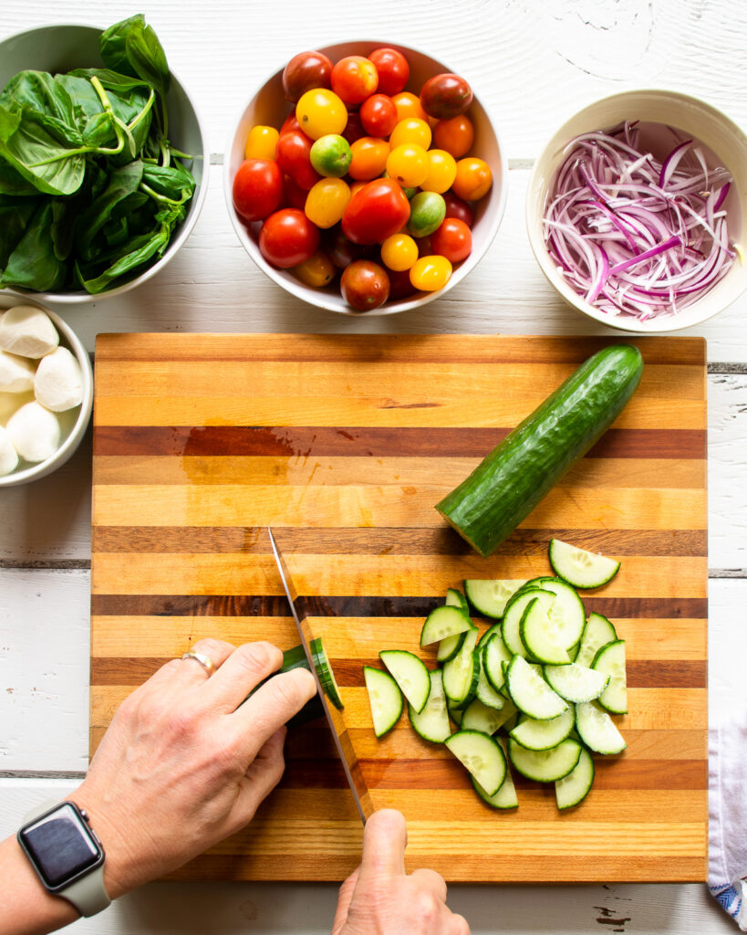 Cucumber being chopped on a wooden cutting board with a bowl of tomatoes, basil, red onion and fresh mozzarella on the side.