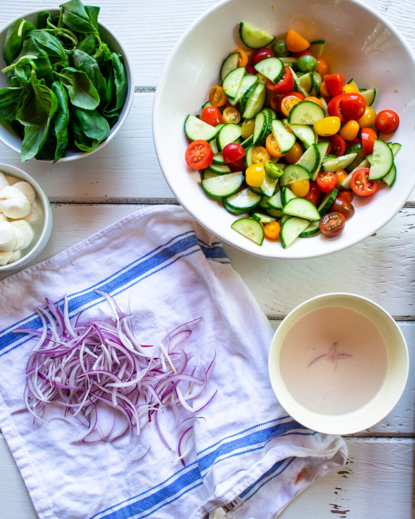 Assembling a tomato cucumber salad on a white table with red onions drying on a kitchen towel.