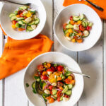 Tomato and Cucumber Salad in a serving bowl with two smaller bowls, orange napkins and forks all on a white table.