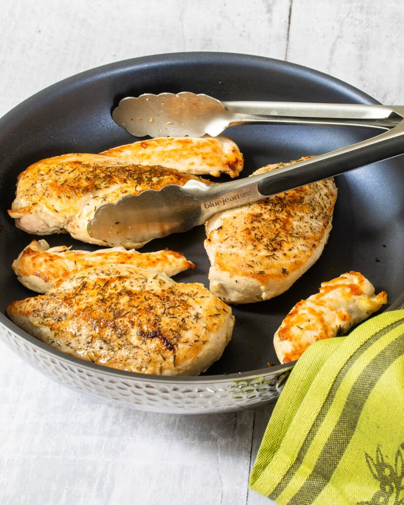 Chicken breasts and tenders in a skillet with a pair of tongs and a green kitchen towel.