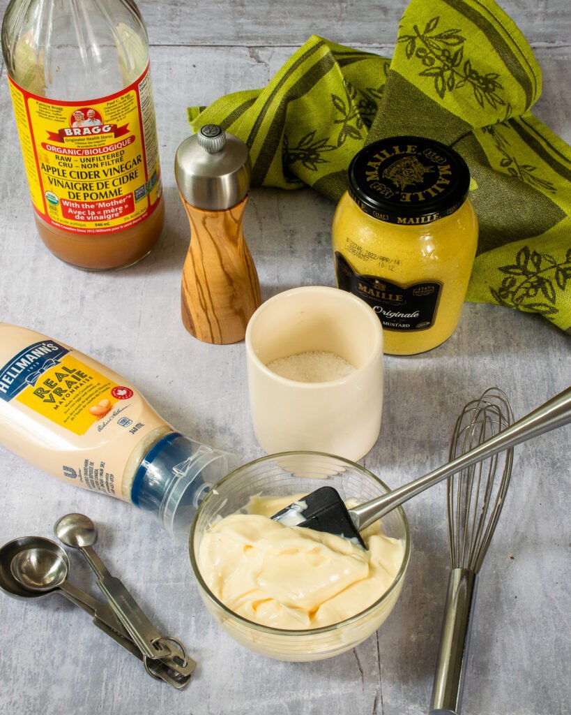 Ingredients on a countertop - mayonnaise, mustard, cider vinegar, salt and pepper, measuring spoons, a whisk and a green kitchen towel.