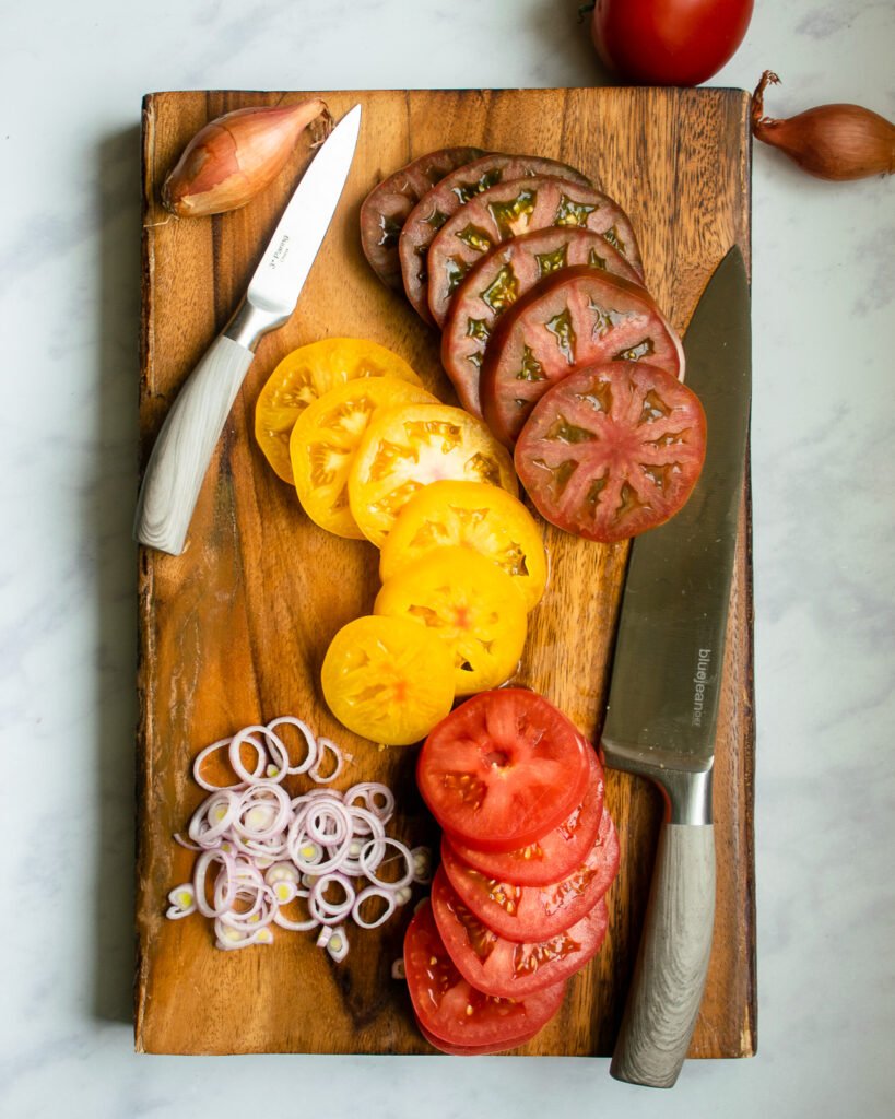 Three different colored tomatoes cut on a wooden cutting board with knives.