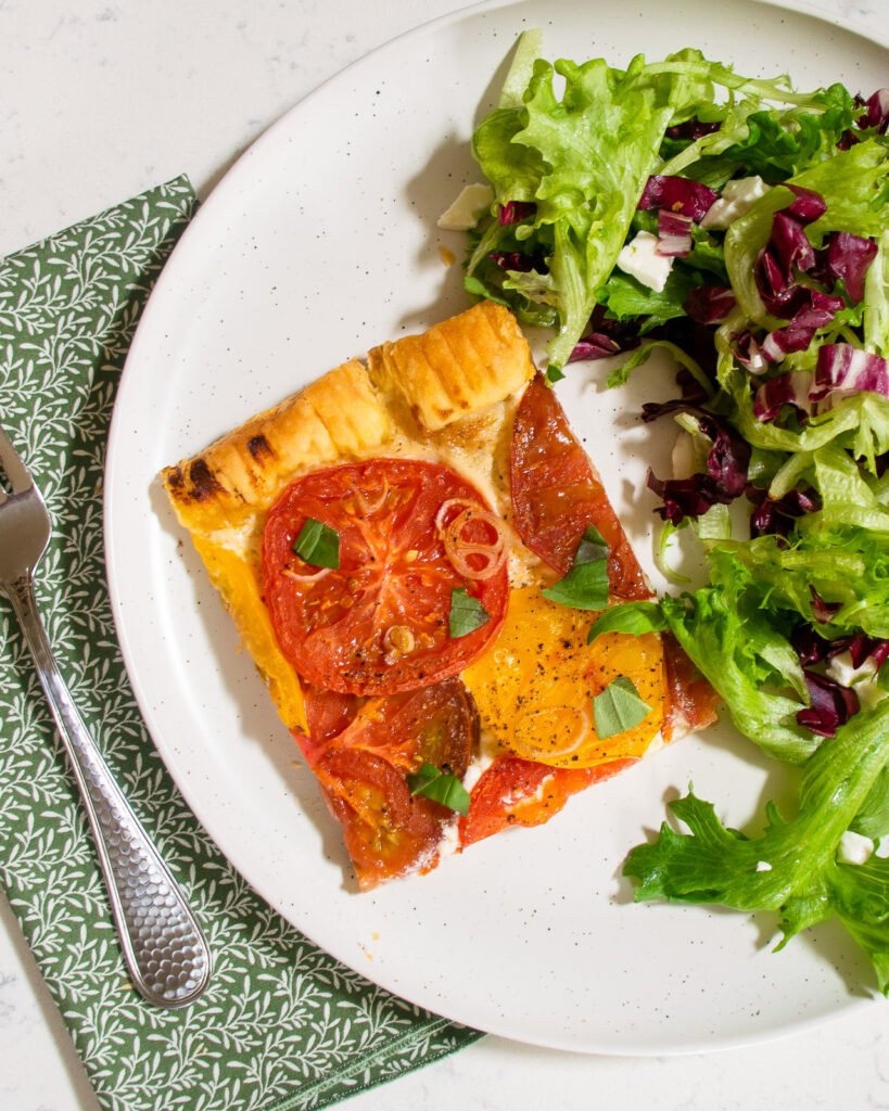 A piece of tomato tart on a white plate with a green napkin and fork.