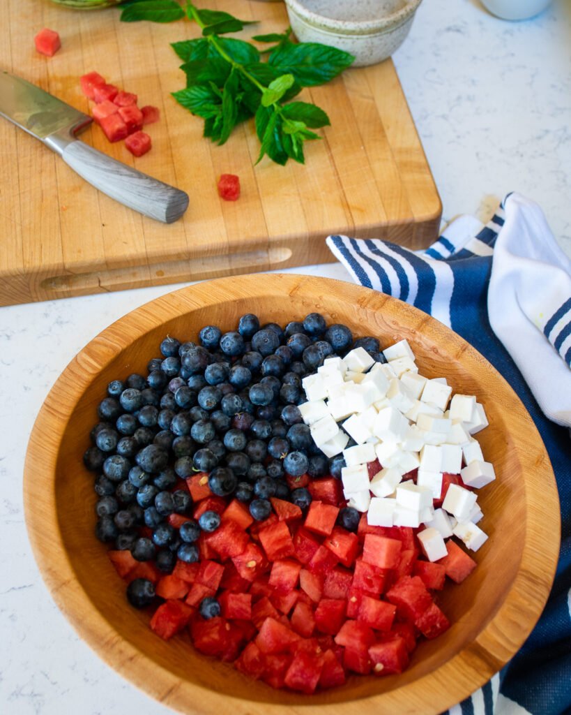 A wooden salad bowl with watermelon, feta and blueberries in it with a cutting board in the background.