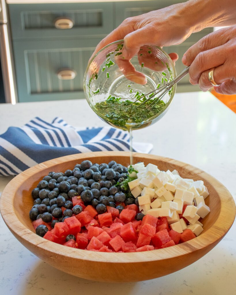 Hands pouring a dressing over a wooden salad bowl with watermelon, feta and blueberries in it.