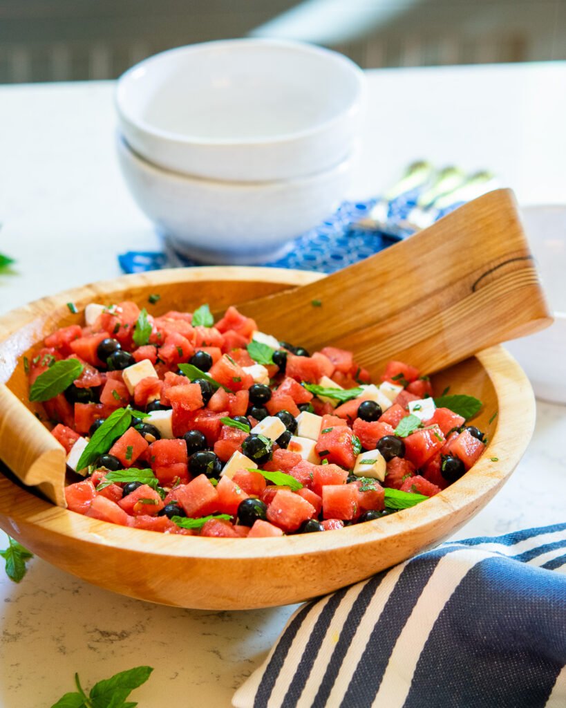 Watermelon feta salad with blueberries in a wooden salad bowl with a blue and white striped towel next to it.