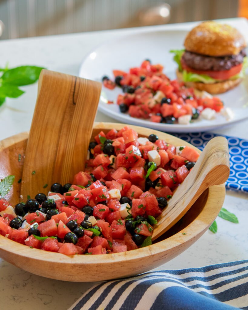 Watermelon feta salad with blueberries in a wooden salad bowl with a blue and a plate with a burger in the background.