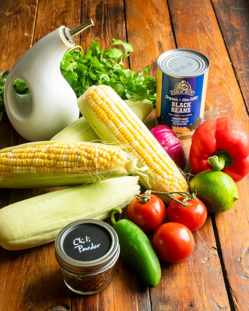 Ingredients on a wooden table - corn, cilantro, peppers, tomatoes, lime, spices and a can of black beans.