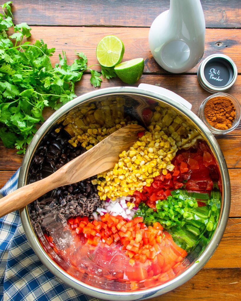 Ingredients in a stainless steel bowl with a wooden spoon, surrounded by a napkin, cilantro, lime halves, olive oil and chili powder.