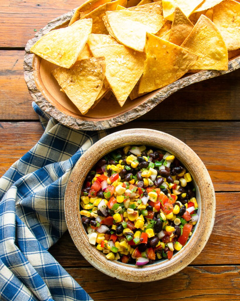 Corn and black bean salsa in a bowl on a wooden table with a blue and white checkered napkin and a wooden bowl of tortilla chips in the background.