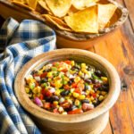 Corn and black bean salsa in a bowl on a wooden table with a blue and white checkered napkin and a wooden bowl of tortilla chips in the background.