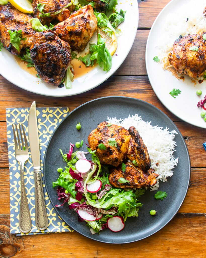 Grilled harissa chicken on a gray dinner plate with a green napkin and cutlery, all on a wooden table.