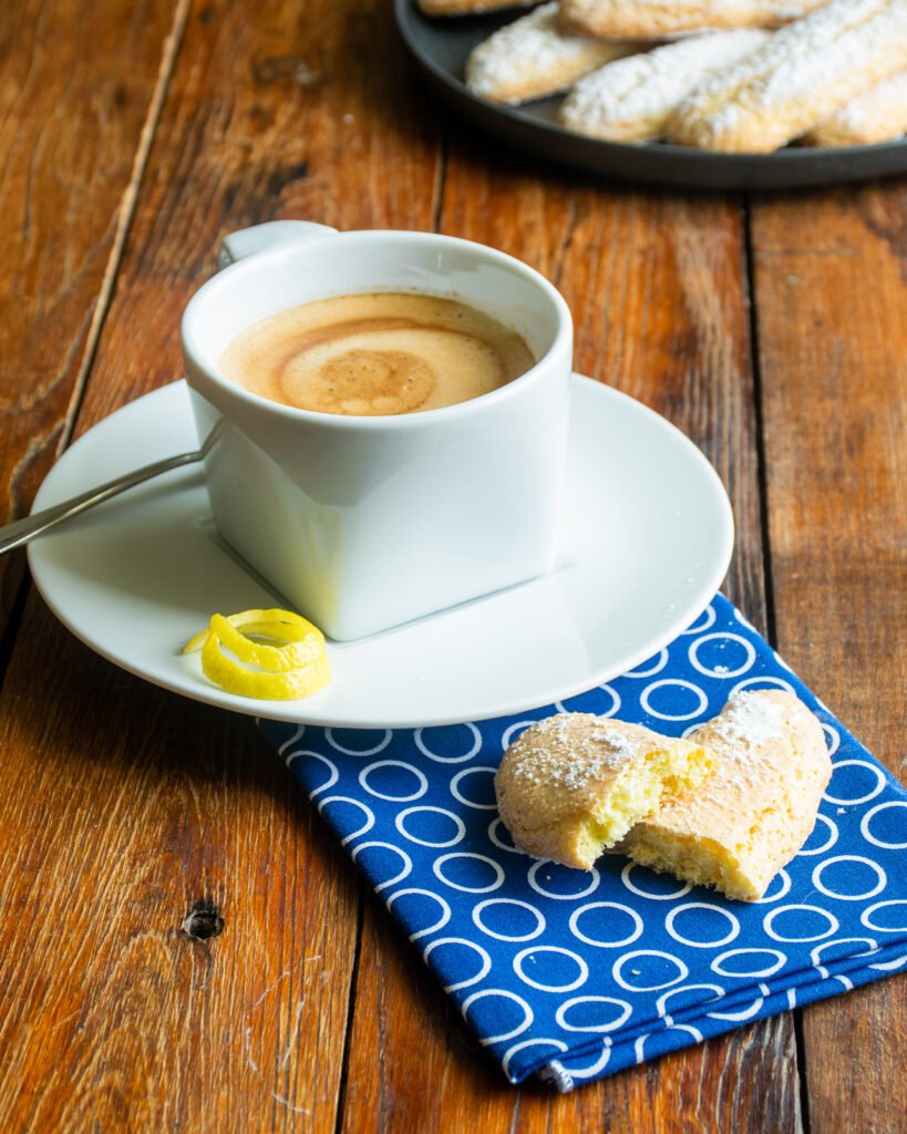 A cup of coffee with a lady finger on the side, along with a lemon peel and a blue polka dot napkin.