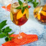A glass of white wine sangria on a red and yellow striped napkin with a pitcher in the background.