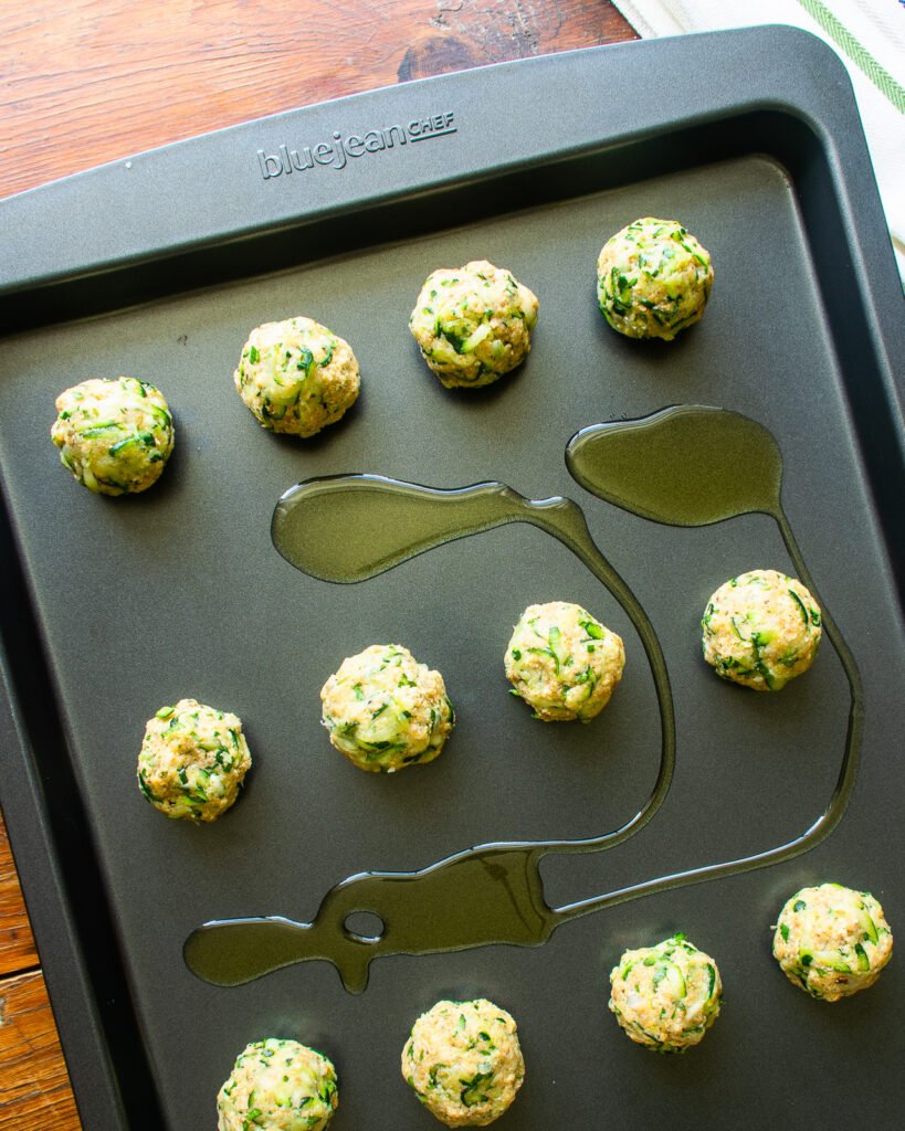 raw zucchini meatballs on a sheet pan with olive oil drizzled on the pan.