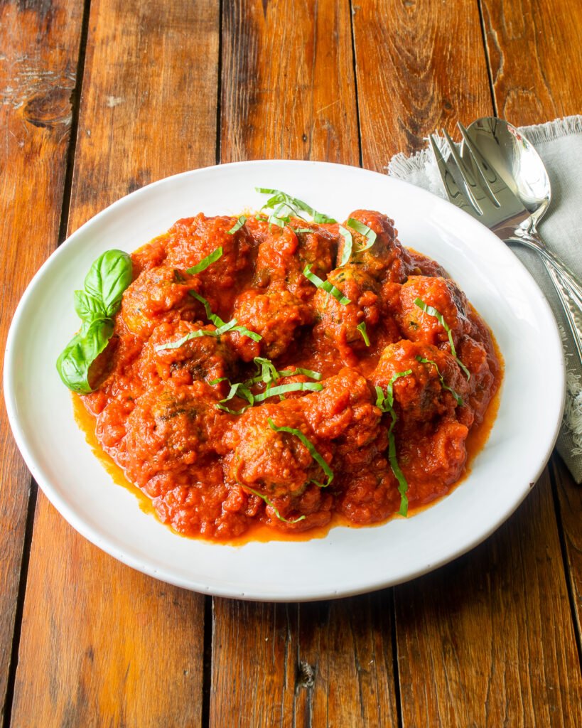 Zucchini meatballs in marinara sauce on a white plate with basil on top and serving utensils on the side.