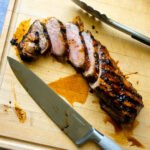 Grilled pork tenderloin sliced on a cutting board with a knife and a pair of tongs.