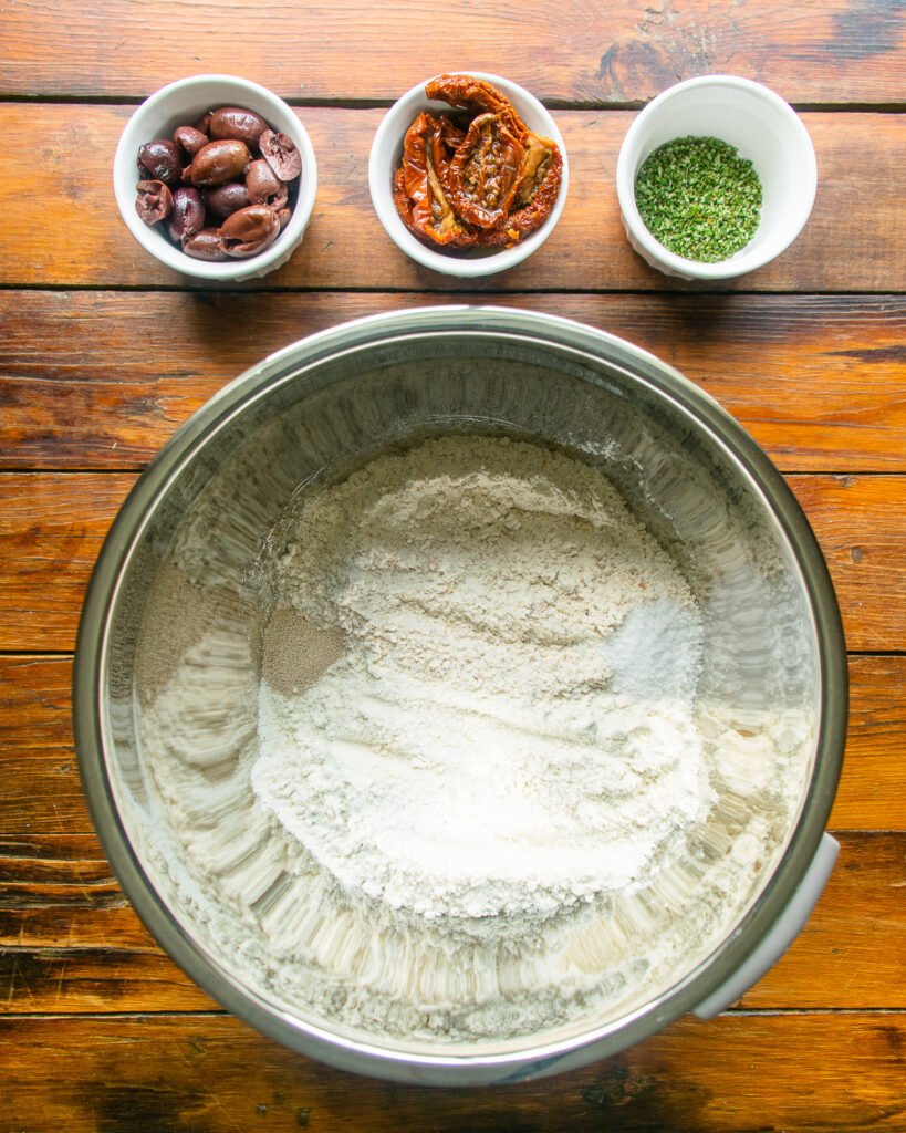 Flour, yeast and salt in a stainless steel bowl with small bowls of olives, sun-dried tomatoes and fresh rosemary next to it.