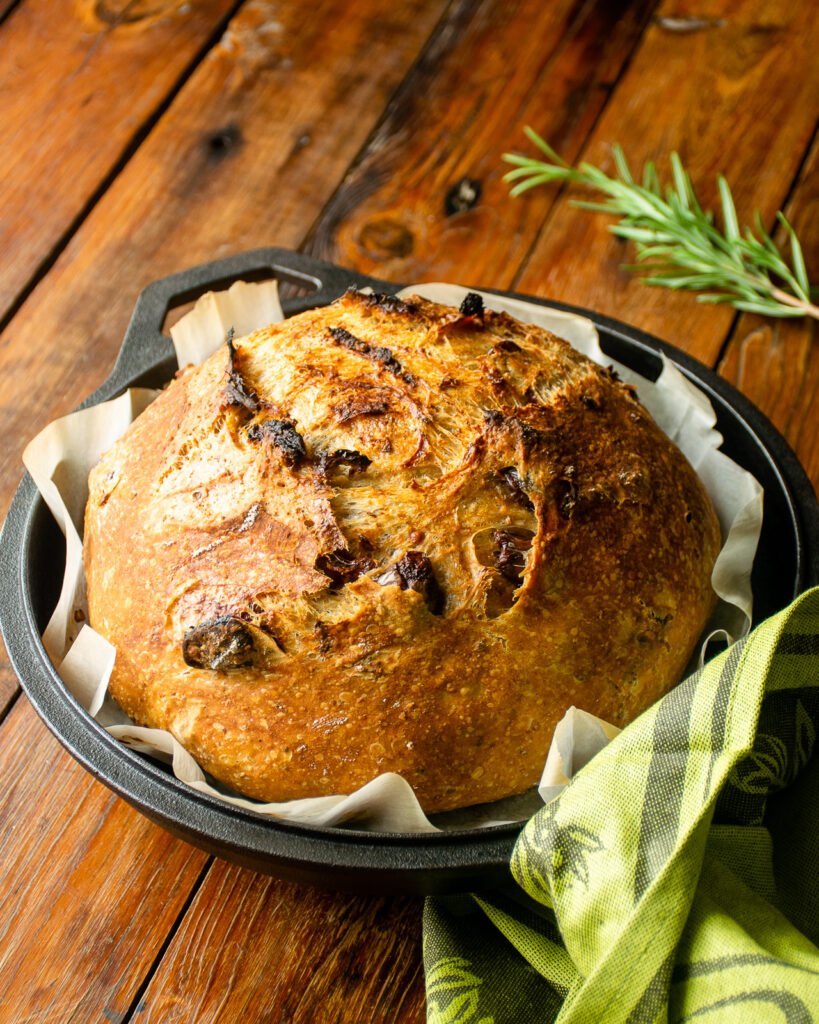 No knead bread with sun-dried tomatoes and olives baked in a cast iron pan, sitting on a wooden table with a green kitchen towel.