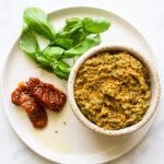 Sun-dried tomato pesto in a small bowl on a white plate with sun-dried tomatoes and fresh basil.