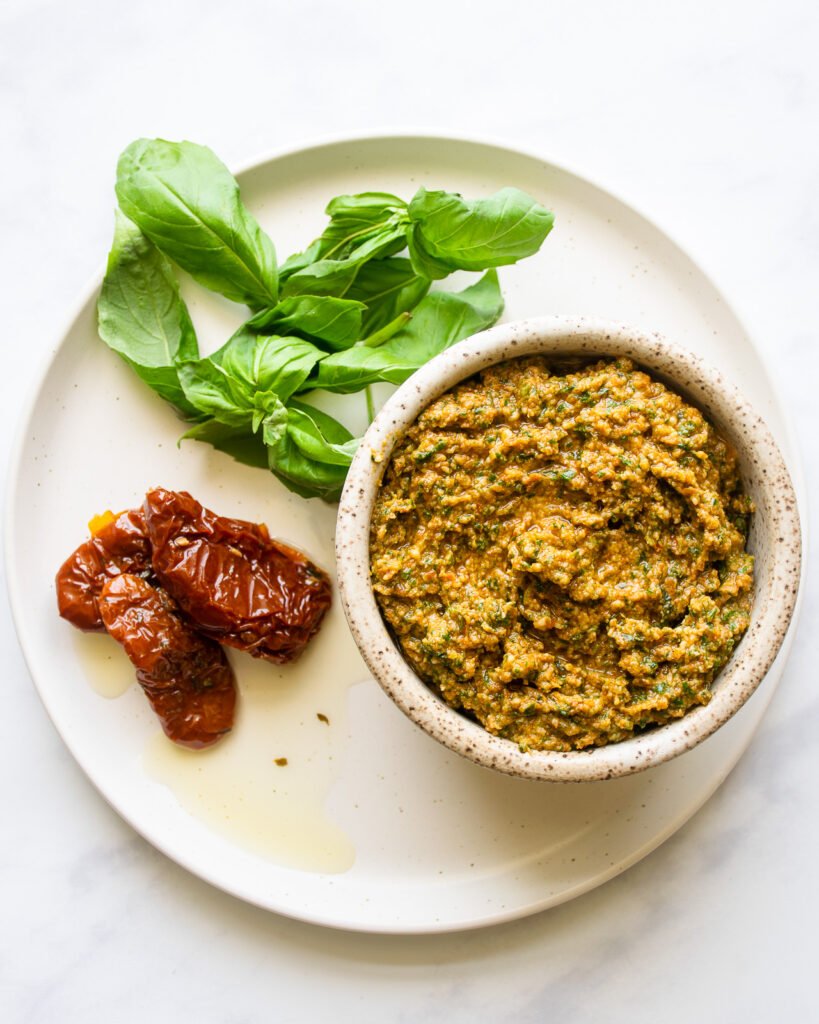 Sun-dried tomato pesto in a small bowl on a white plate with sun-dried tomatoes and fresh basil.